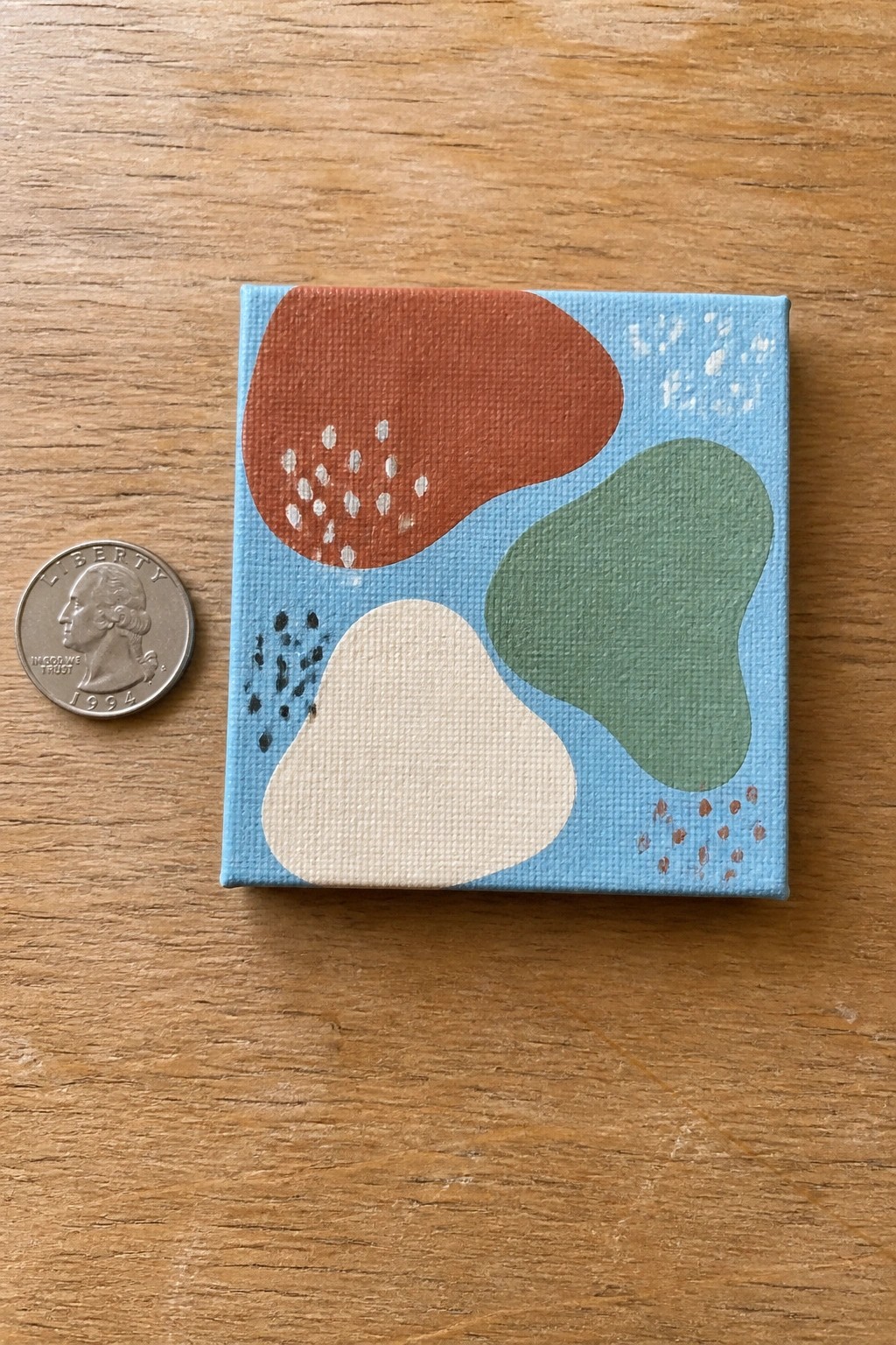 Miniature abstract canvas with orange, green, beige shapes beside a US quarter on wood