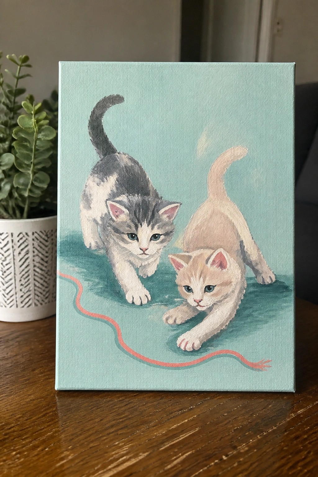 Painting of two kittens on light blue background playing with red string.