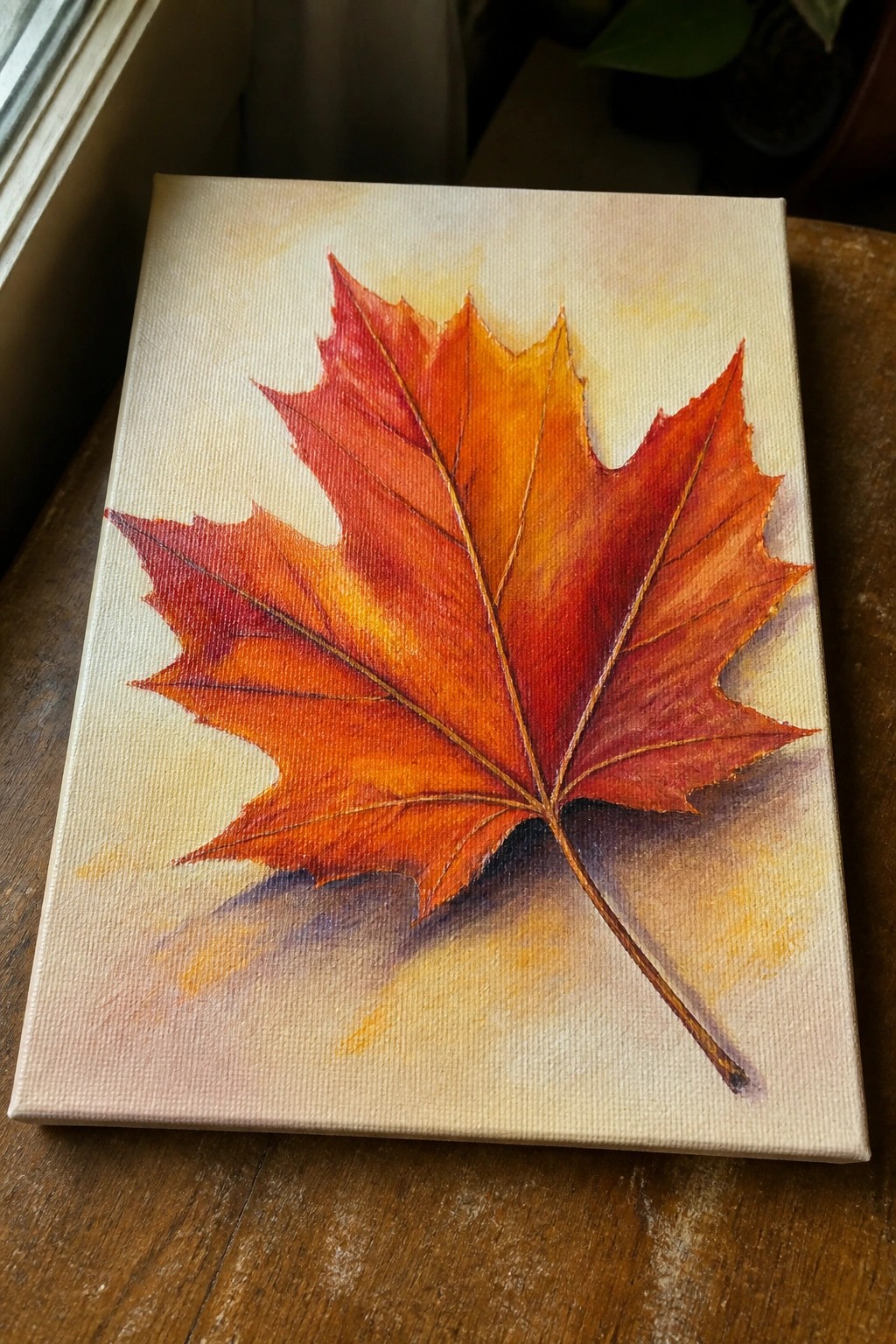 Vibrant red-orange maple leaf painting on white canvas atop wooden table