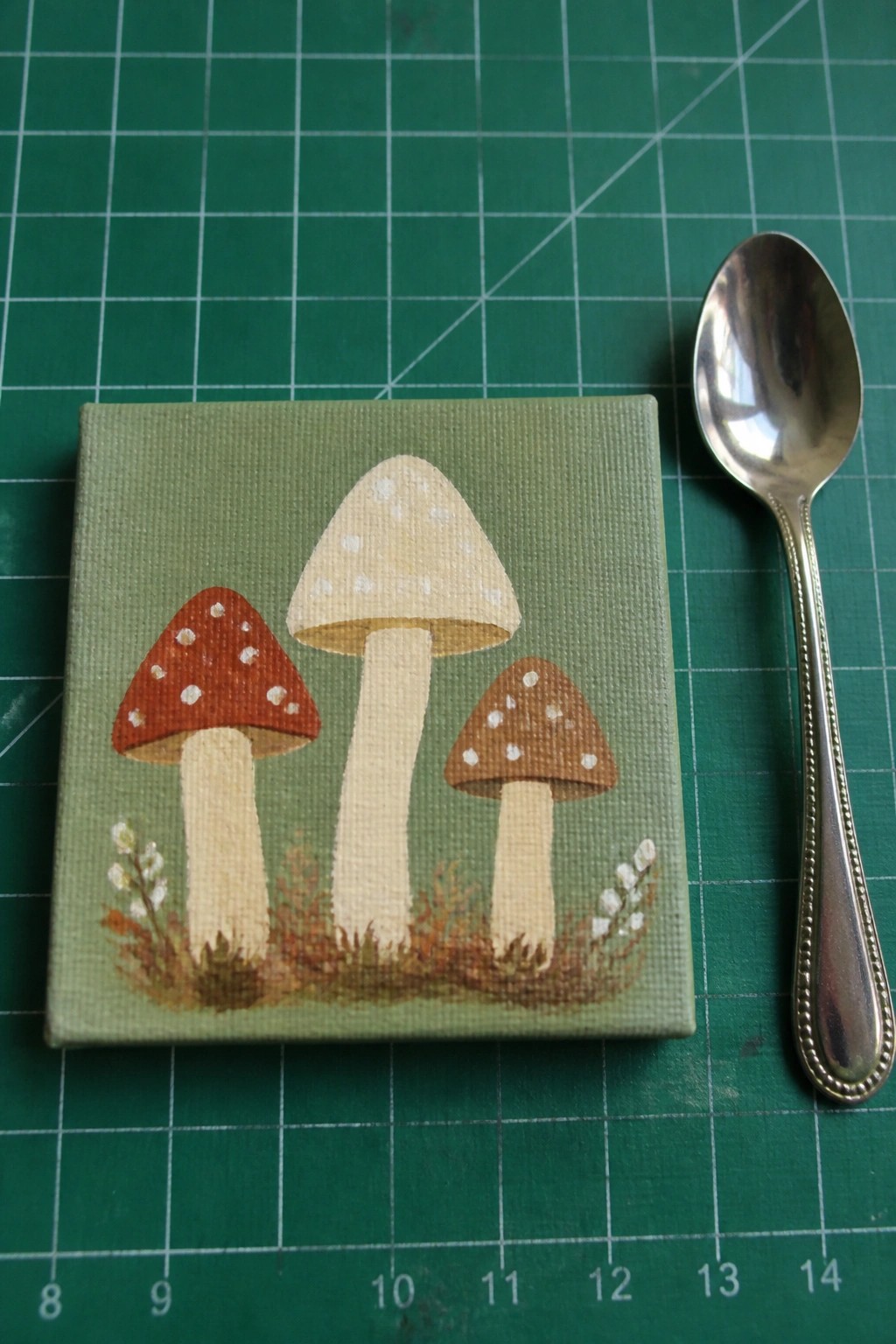 Tiny canvas painting of three spotted mushrooms beside silver spoon on green mat
