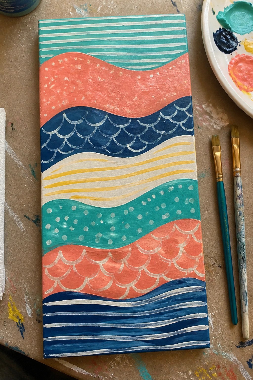 Wavy abstract canvas painting in teal coral blue with scalloped patterns beside brushes and palette.