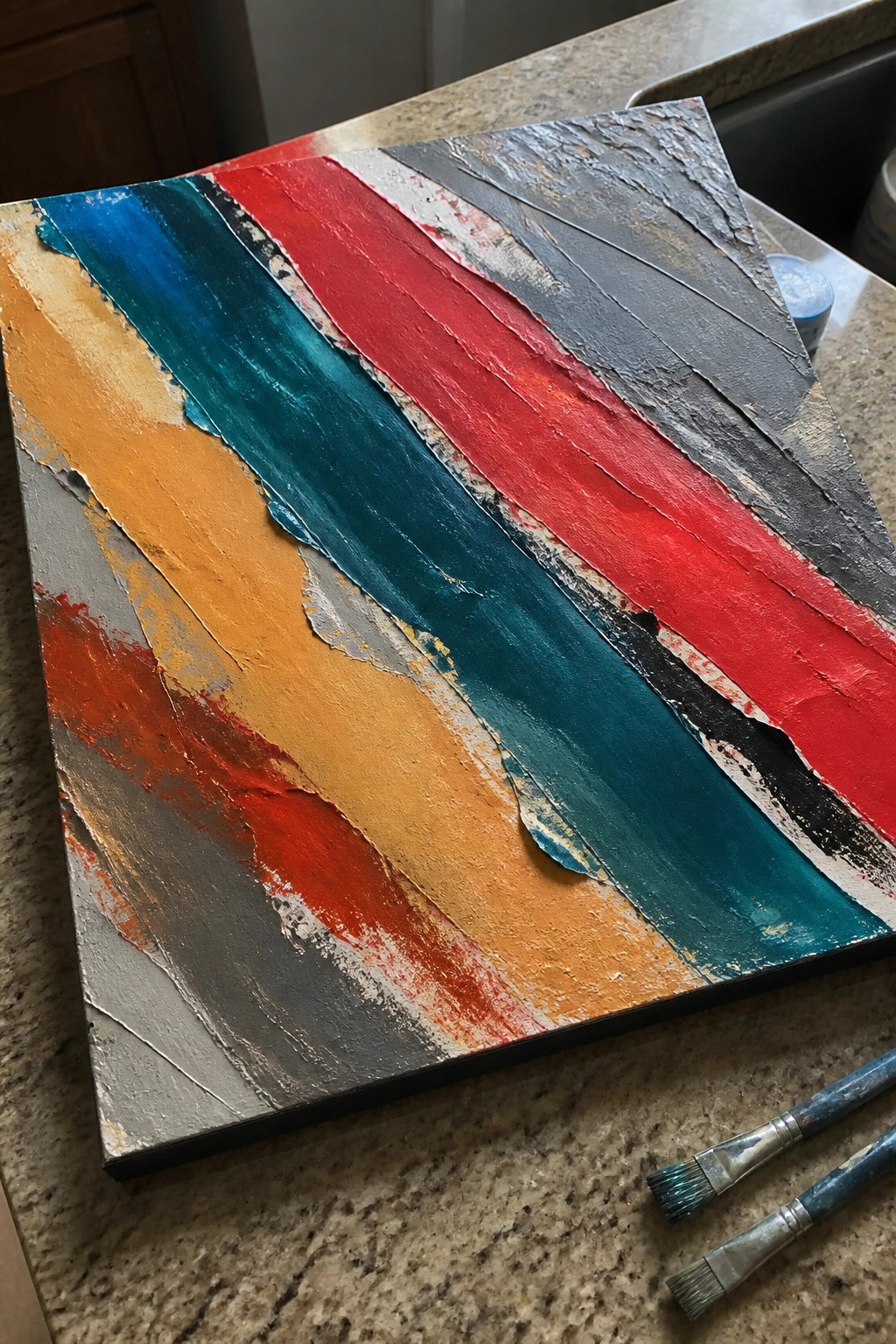 Abstract painting with thick textured strokes of red, teal, orange, and gray on canvas near brushes.