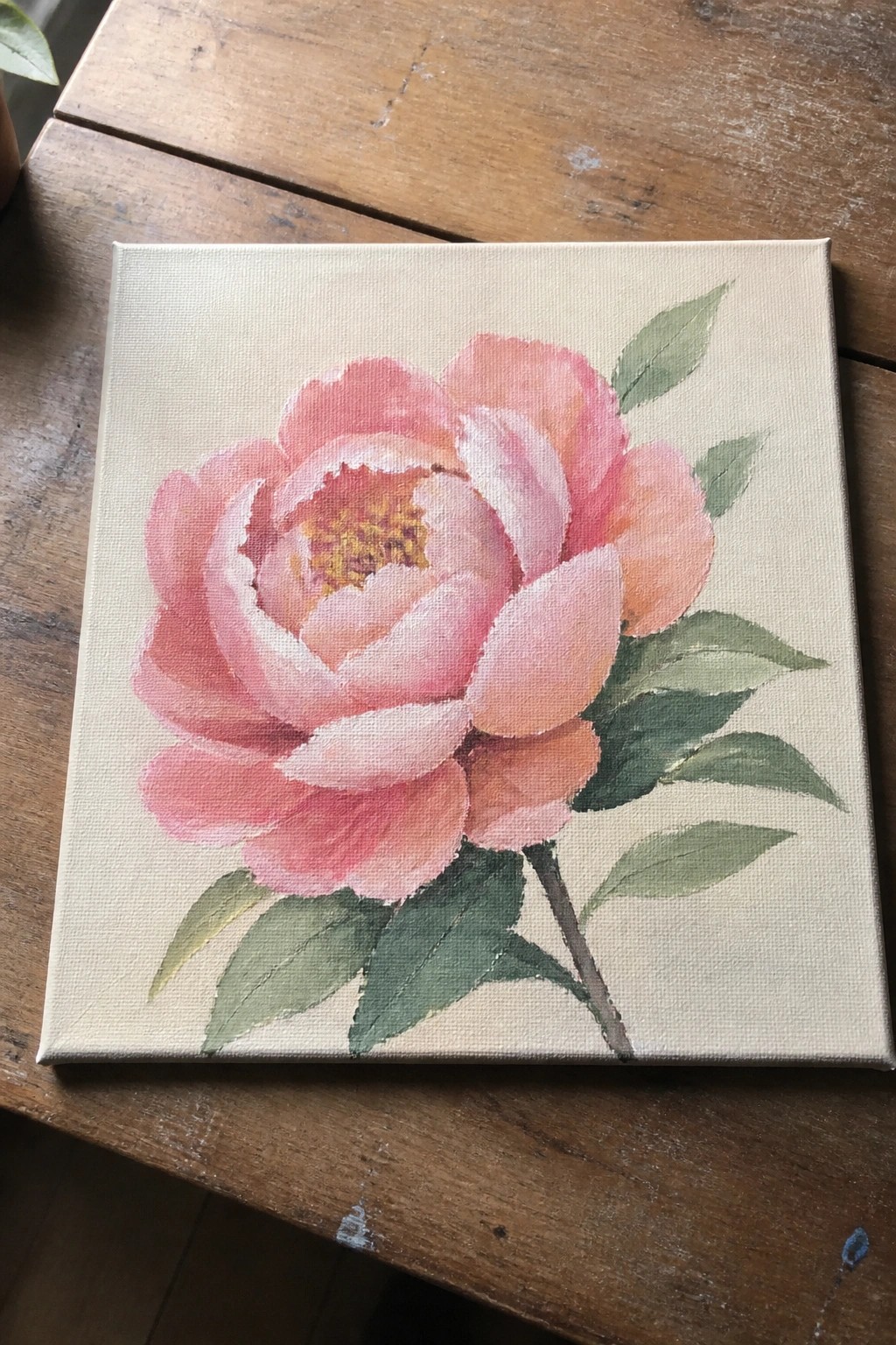 Pink peony painting with green leaves on canvas atop wooden table