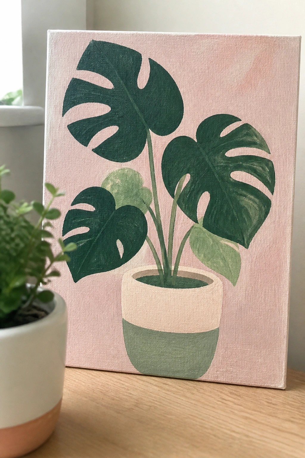 Painting of monstera plant in two-tone pot on pink canvas with real plant nearby