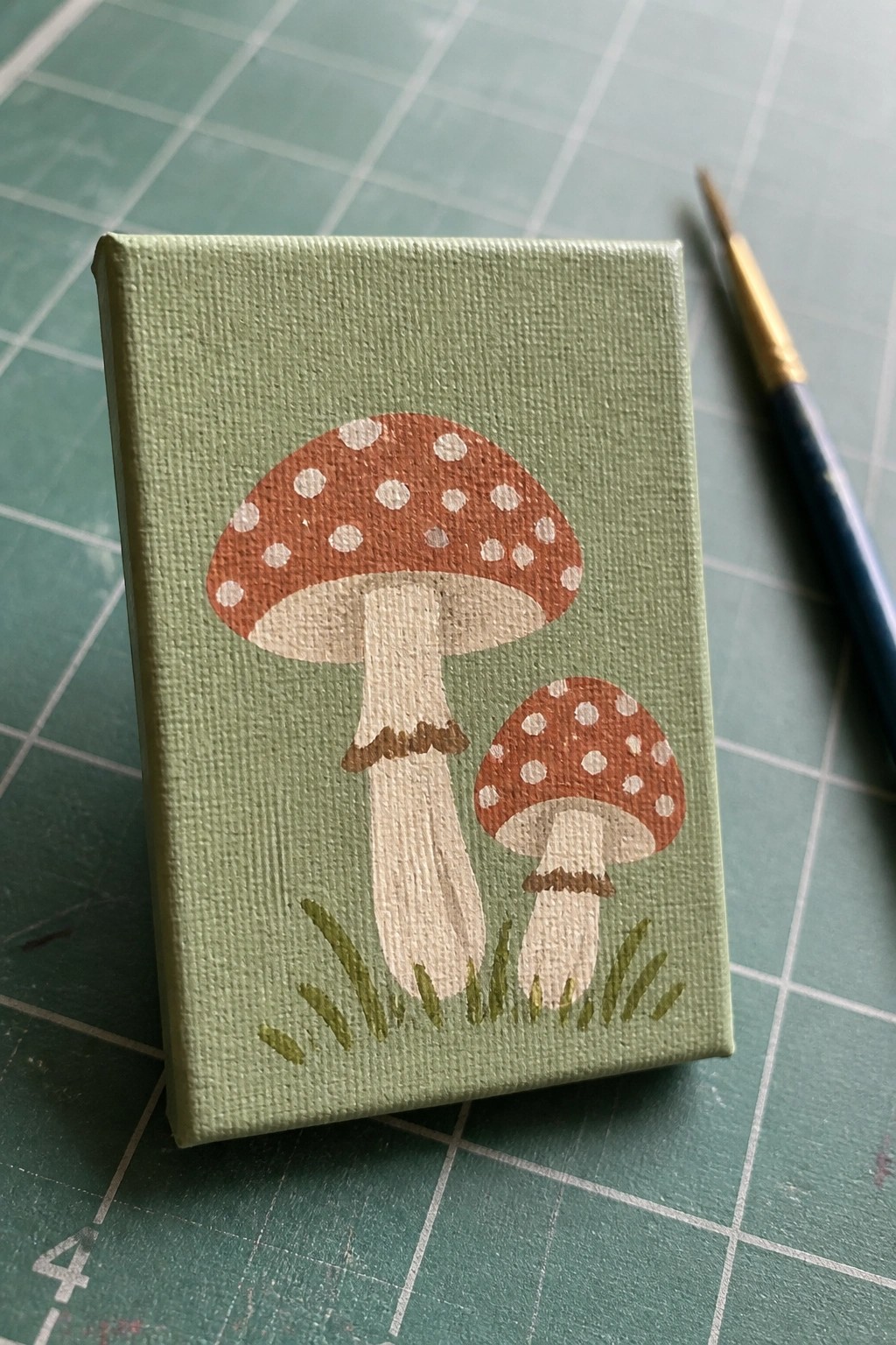 Mini canvas with two red-spotted mushrooms and paintbrush on green mat