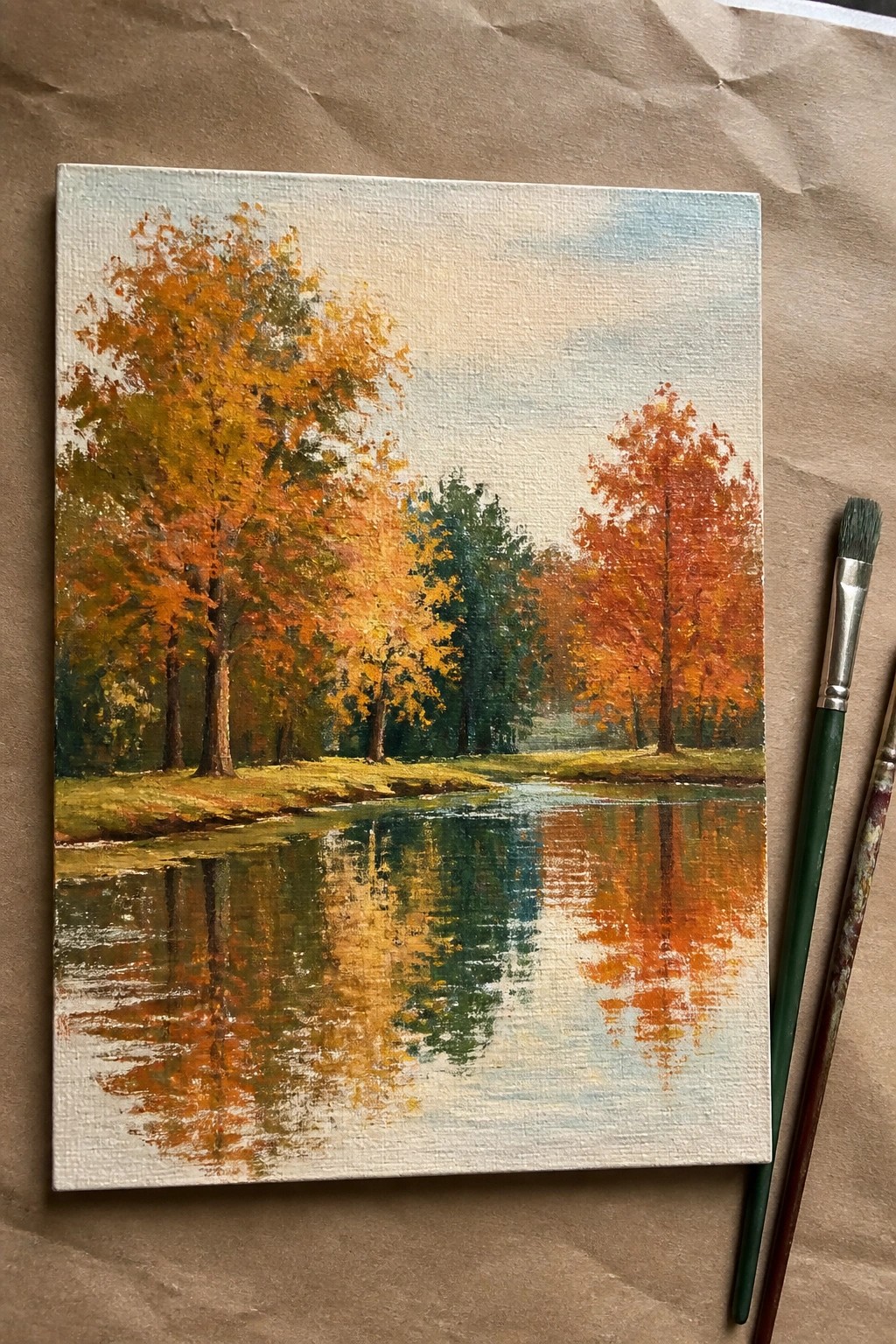 Autumn landscape painting of orange trees reflected in river beside paintbrushes