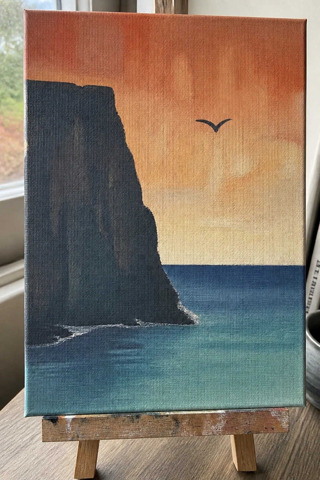 Painting on easel of dark cliff beside blue-green sea under orange sunset sky with bird.