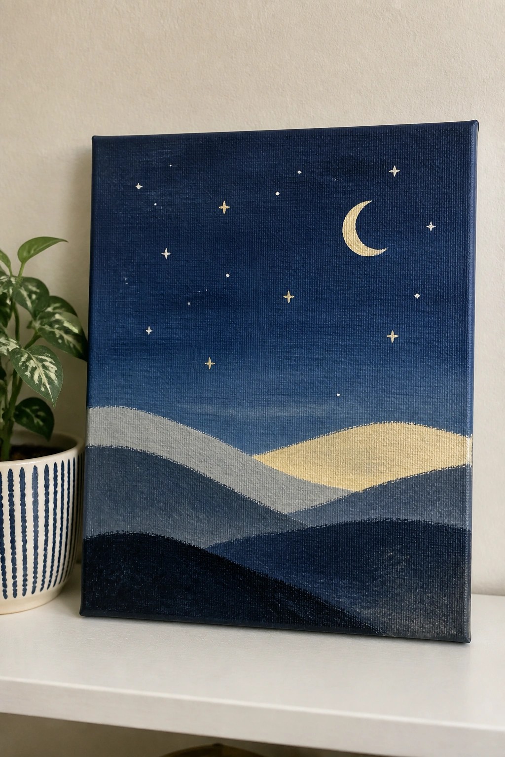 Canvas painting of crescent moon and stars above layered blue hills beside potted plant.