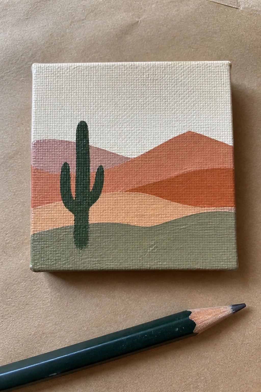 Tiny canvas painting of cactus in layered orange desert hills beside green pencil