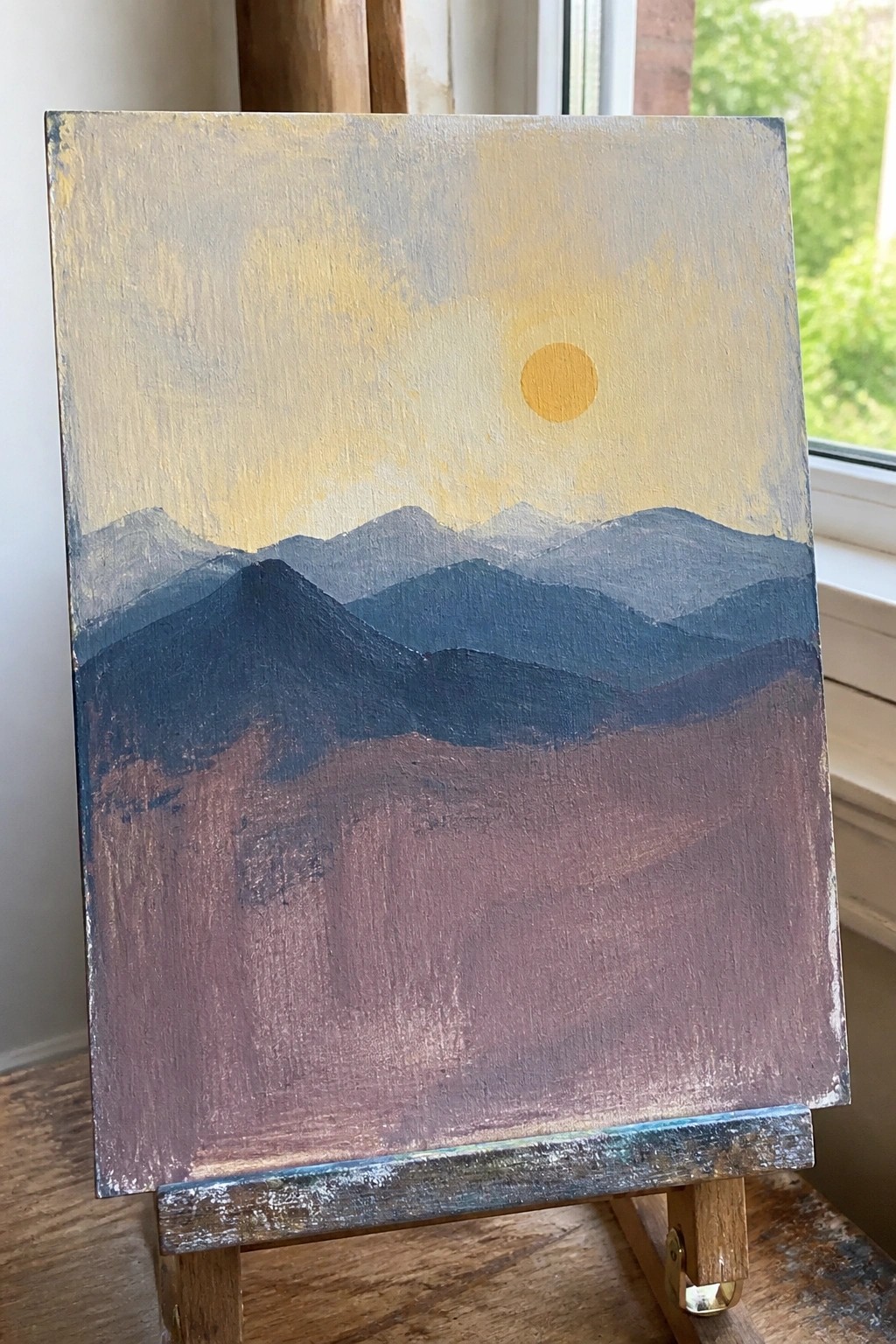 Painting on easel shows layered blue mountains under yellow sun with brown foreground.