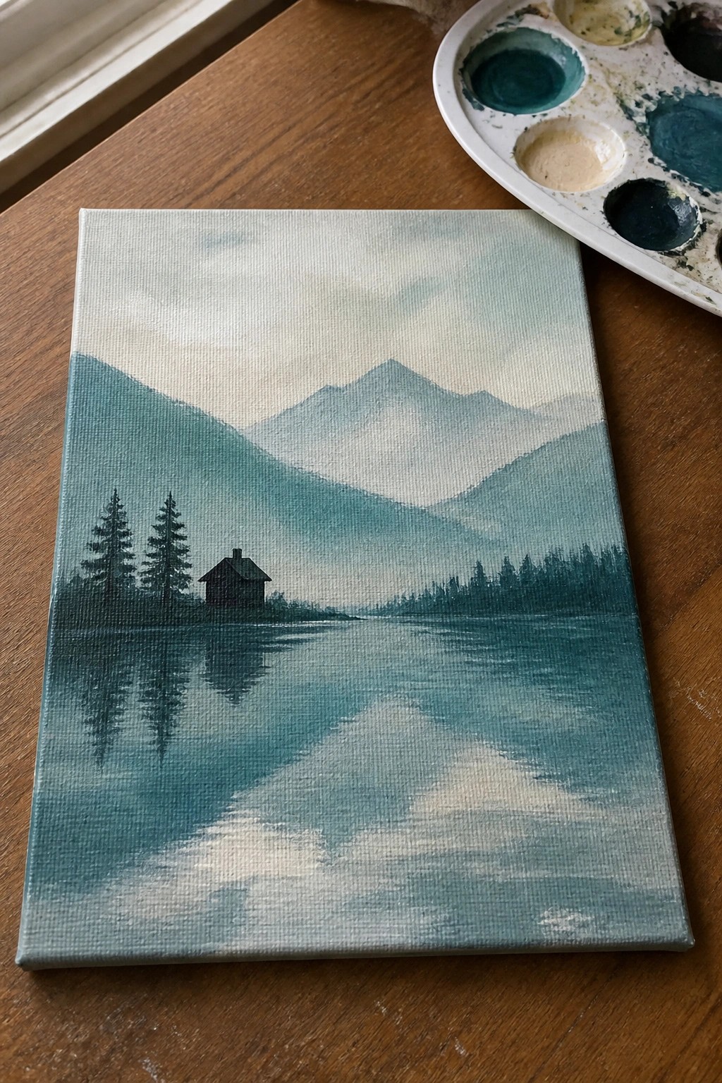 Misty mountain lake watercolor painting on canvas beside paint palette on wooden table