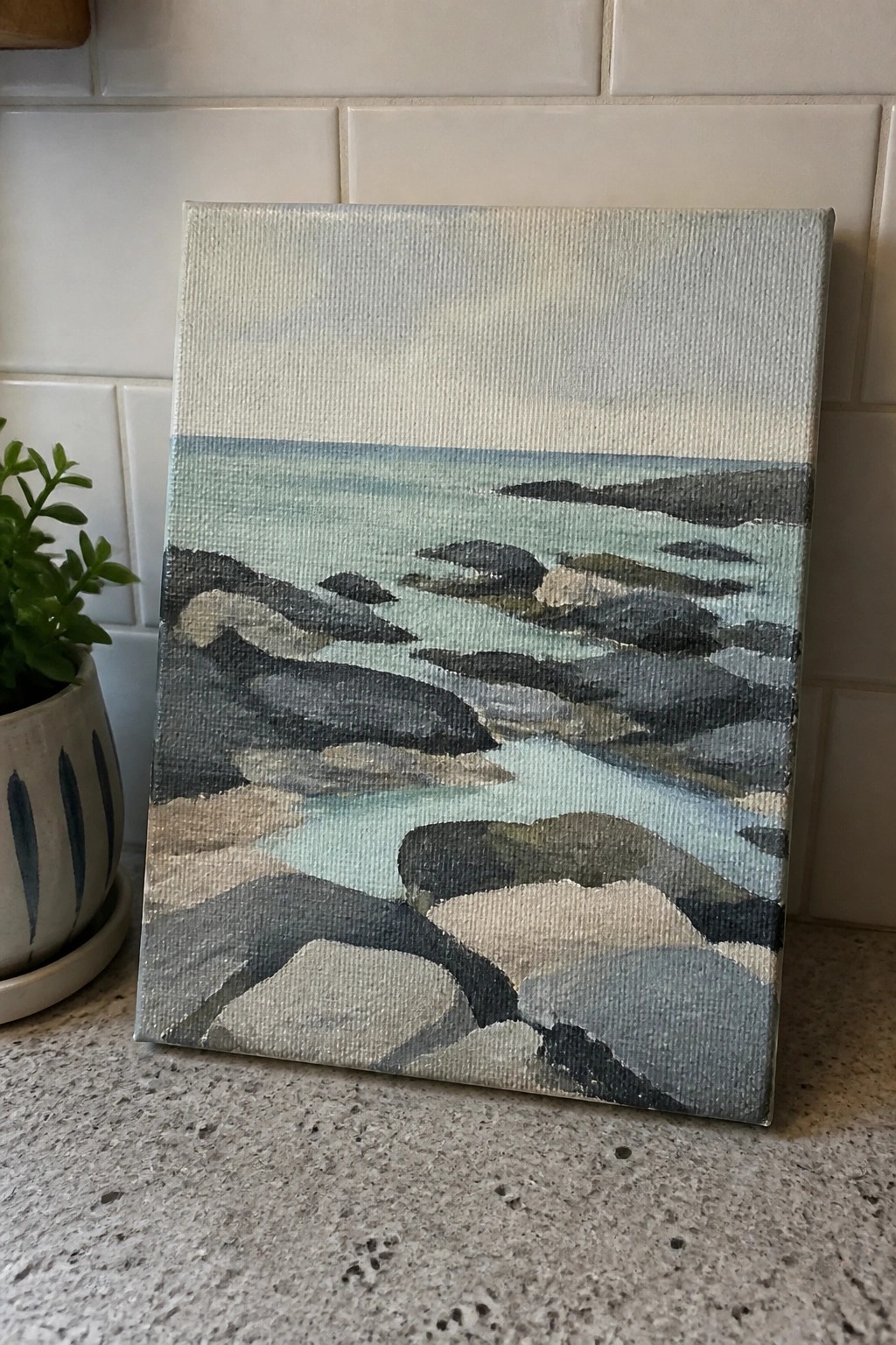 Canvas painting of dark rocks in turquoise water beside potted plant