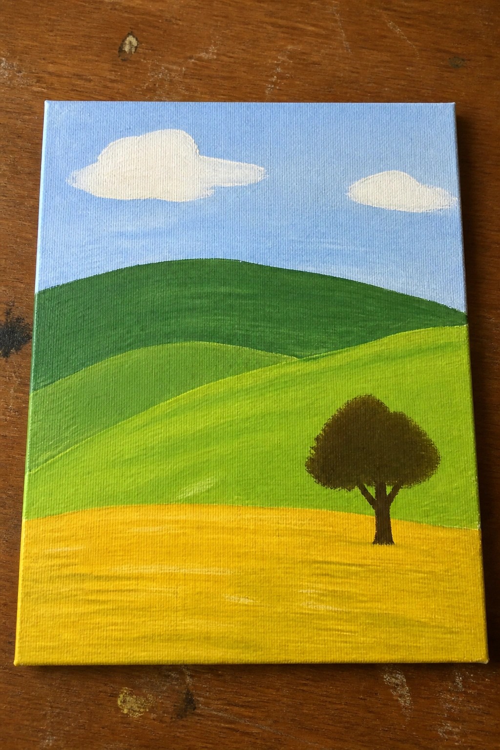 Landscape painting with green rolling hills, yellow field, lone tree, blue sky and clouds.