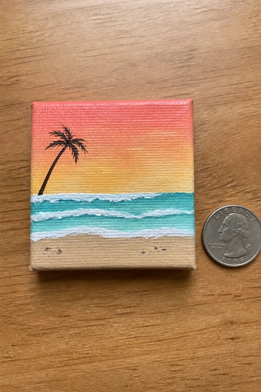 Miniature canvas beach sunset painting with palm tree, waves, and sand beside quarter coin