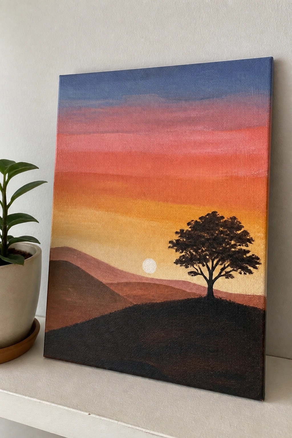 Canvas sunset painting with gradient sky, silhouetted tree on hills, beside potted plant.