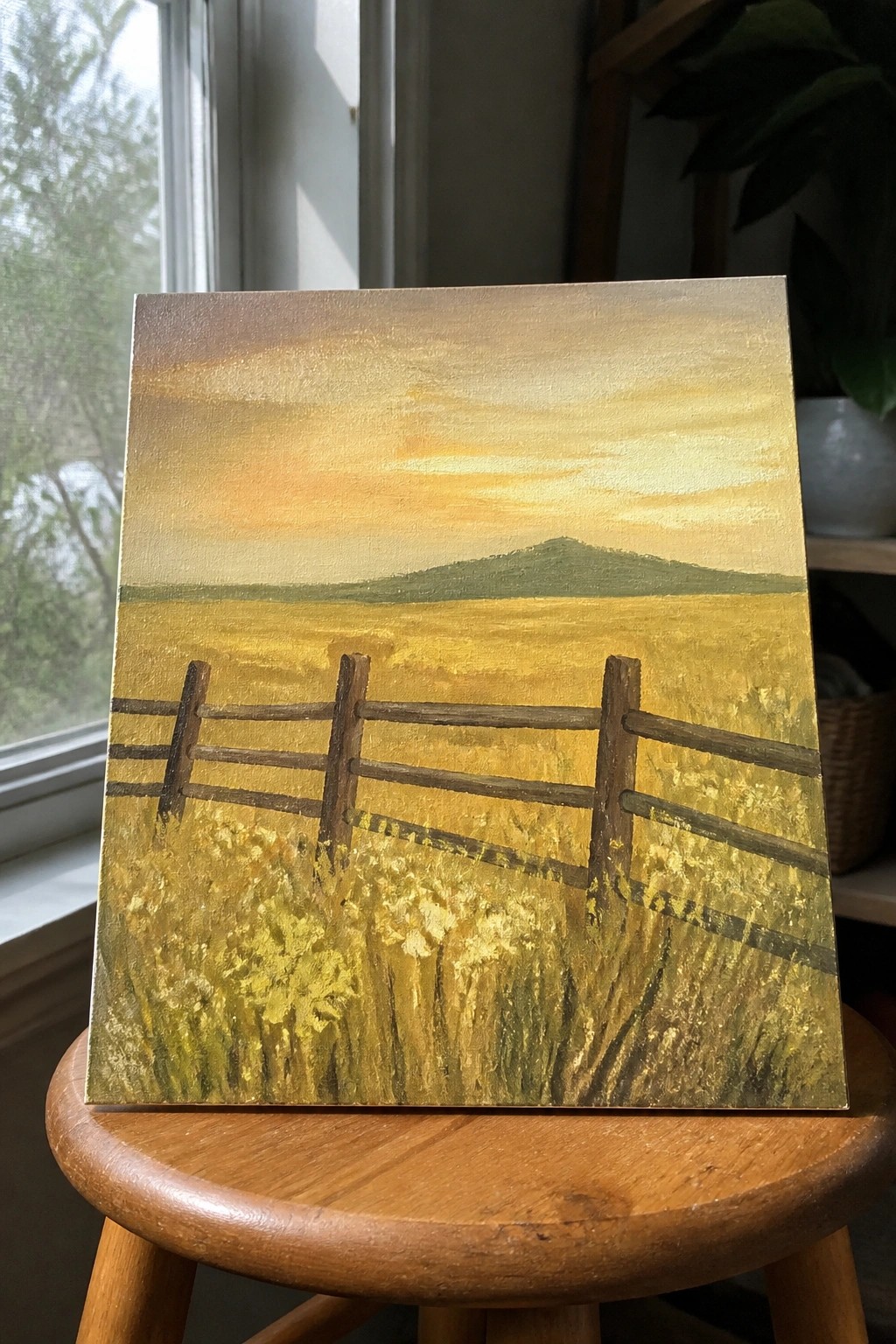 Acrylic painting of a yellow field with a wooden fence, distant green hill, and warm sunset sky resting on a wooden stool.