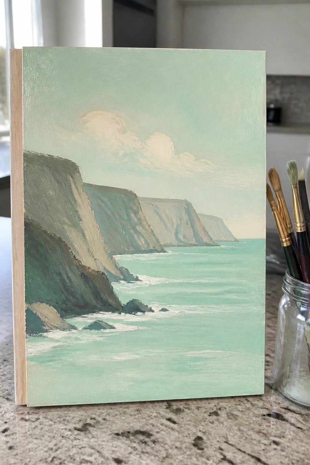 Painting of green cliffs by turquoise sea on canvas beside paintbrushes in jar.