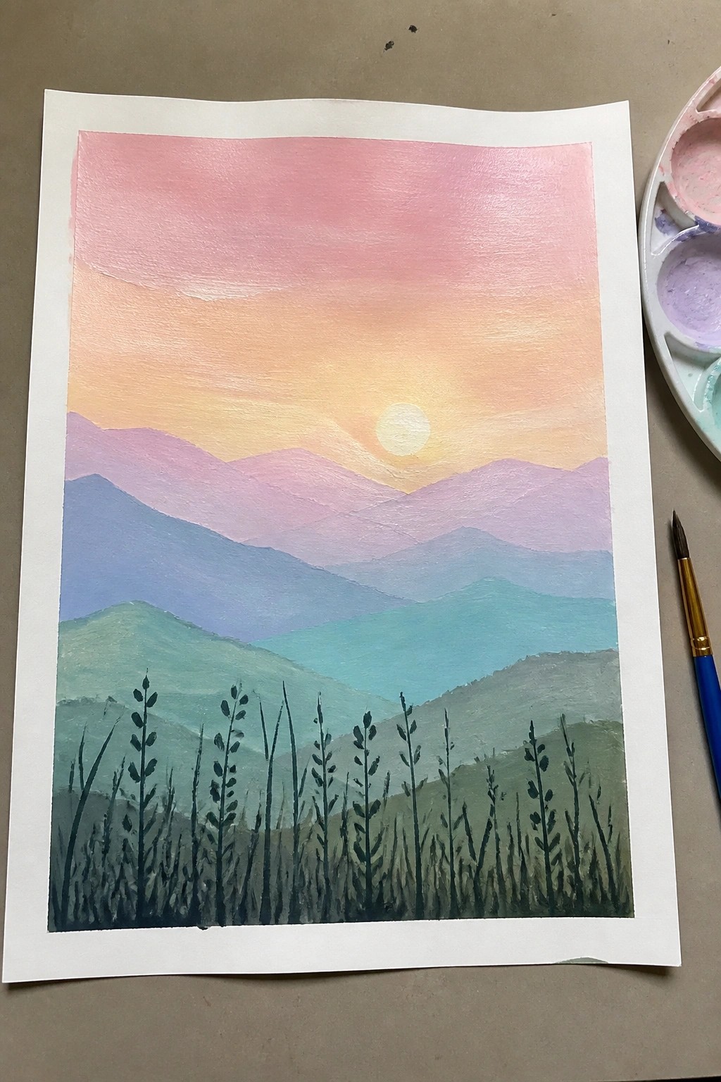 Watercolor sunset landscape with pink-orange sky, layered mountains, sun, and foreground grass beside palette