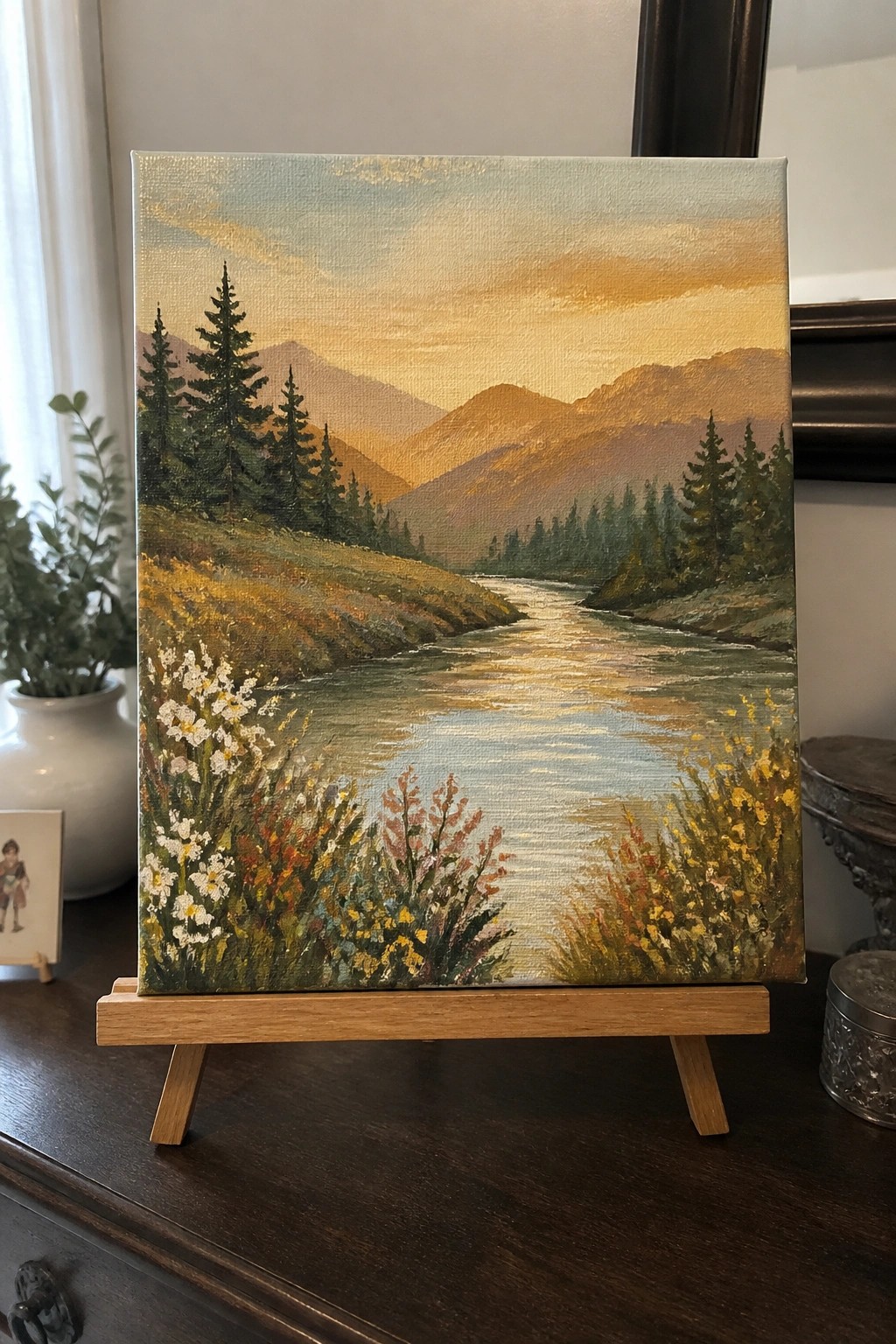 Sunset river landscape painting with mountains, pine trees, and colorful wildflowers on easel.