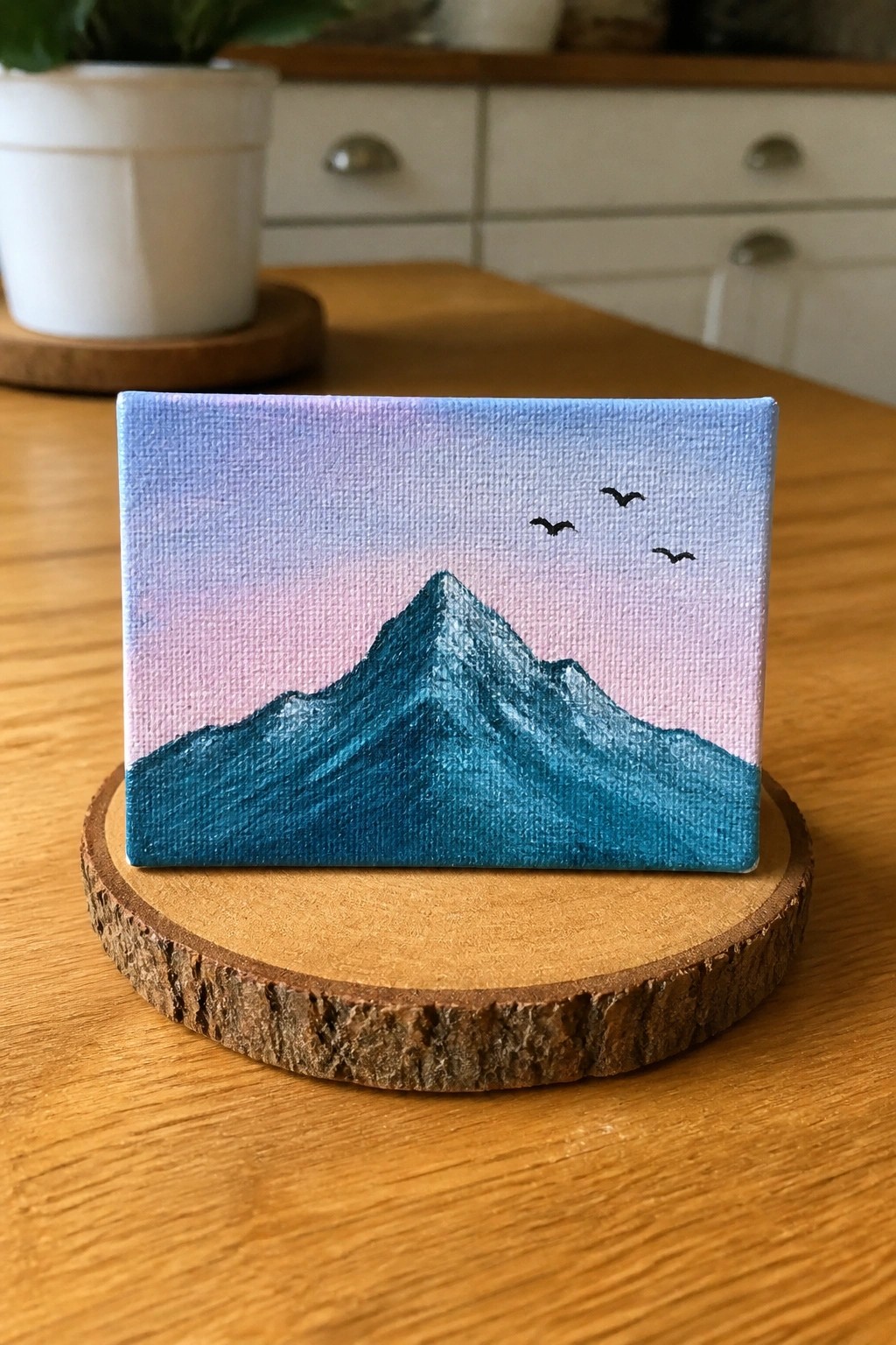 A small acrylic painting of a teal mountain under a blue to pink gradient sky with three black birds, displayed on a wooden slice.
