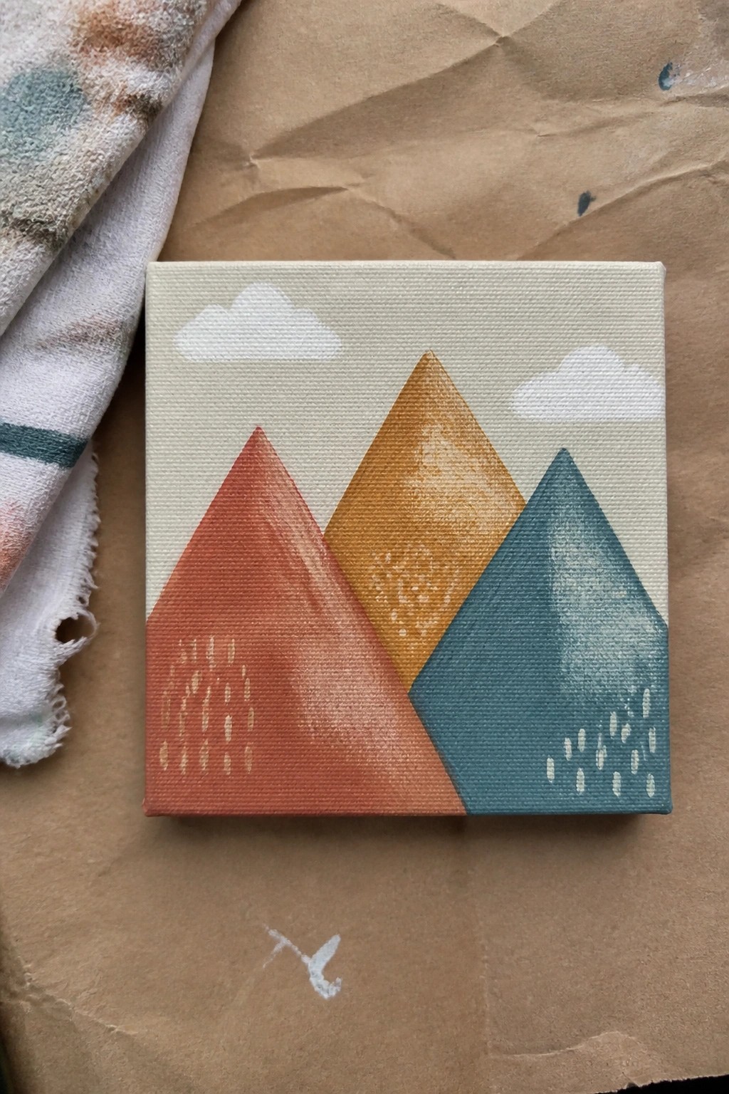 Small canvas with red, orange, and teal mountains, clouds, and rain beside fabric
