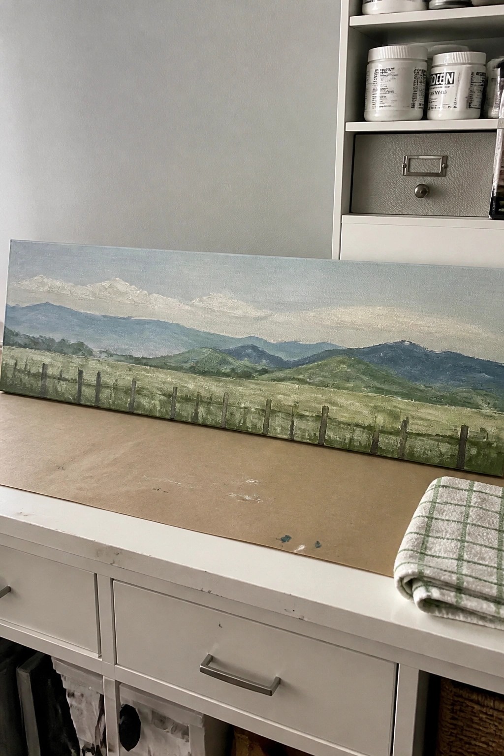 Landscape painting of green fields, fence, and mountains on white cabinet with checkered cloth