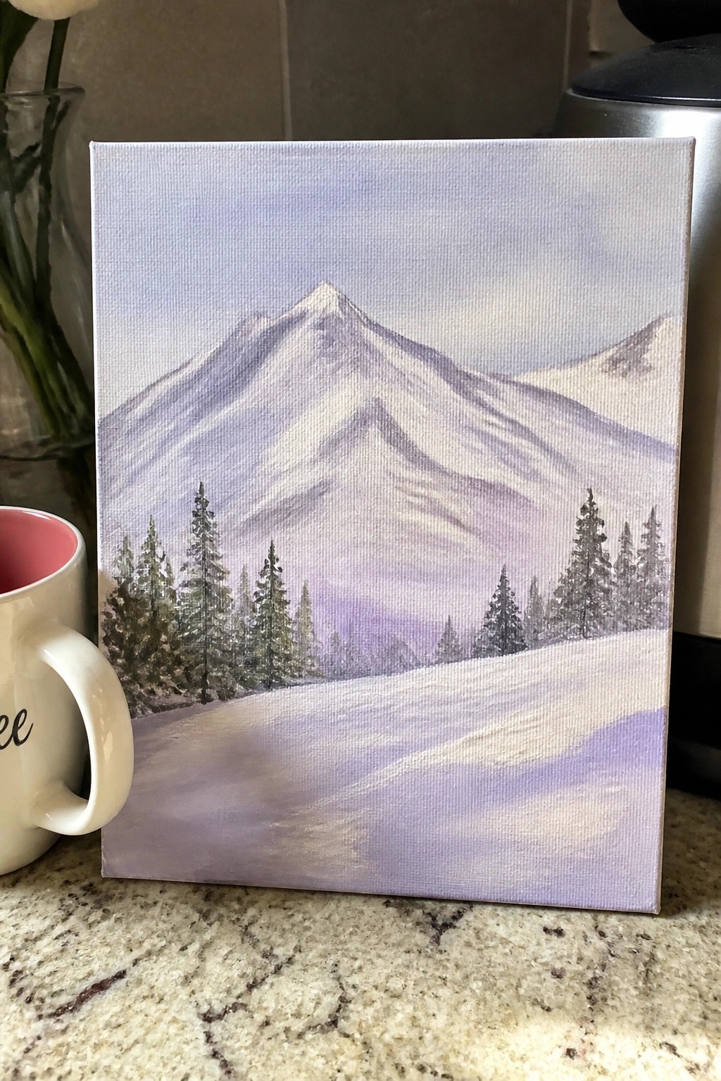 Purple-hued painting of snowy mountains and pine trees on canvas beside white mug