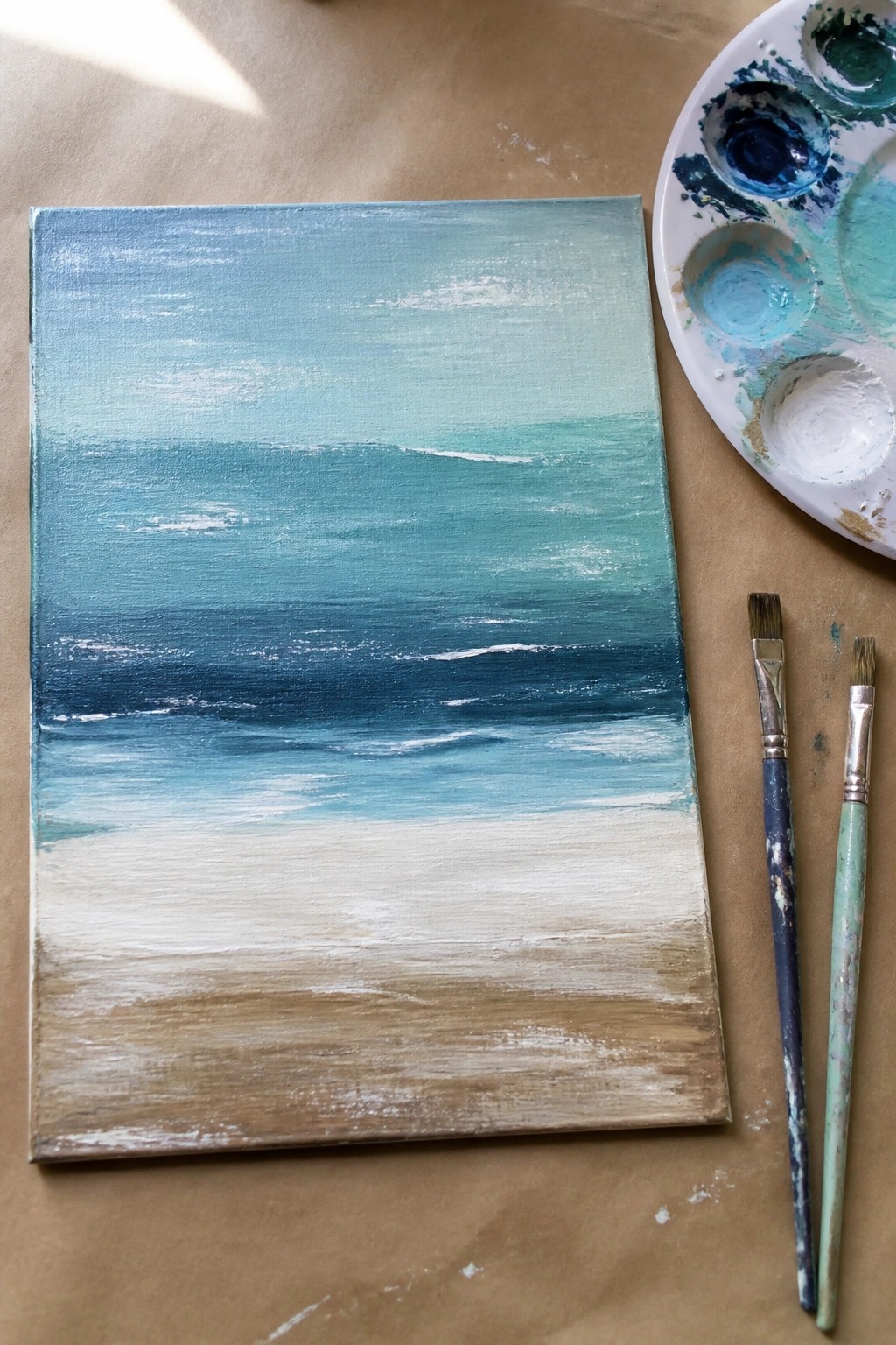 Abstract seascape painting in blues and sands beside palette and brushes