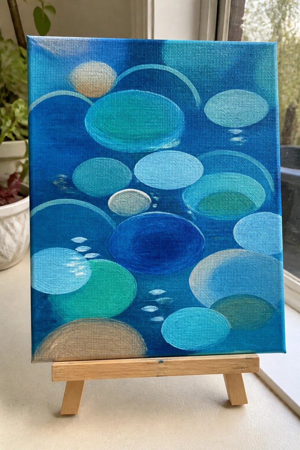 An acrylic painting of overlapping blue, teal, and green circles on a blue background displayed on a small wooden easel.