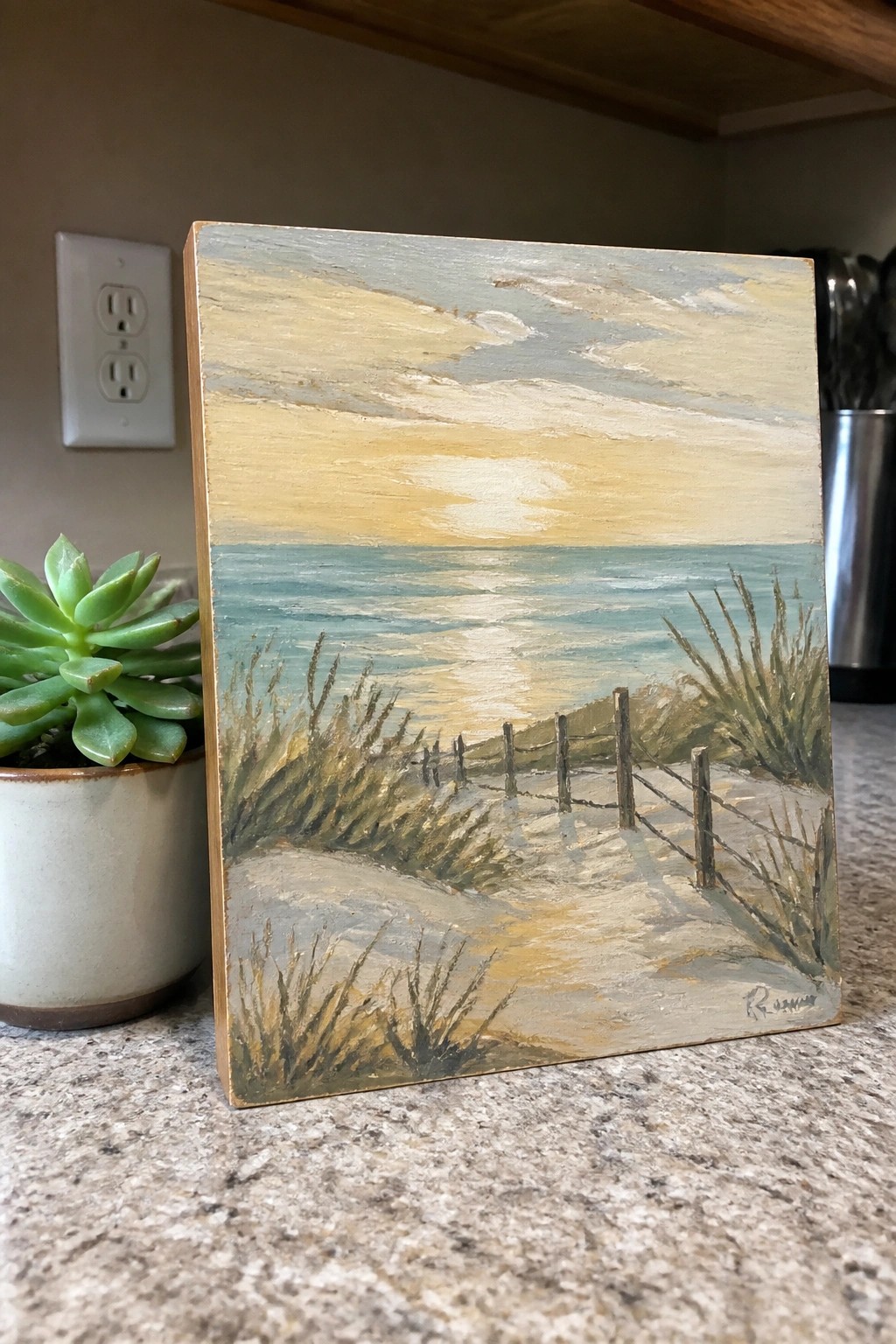 Beach sunset painting with sandy path, fence, ocean waves, and grass beside potted succulent.