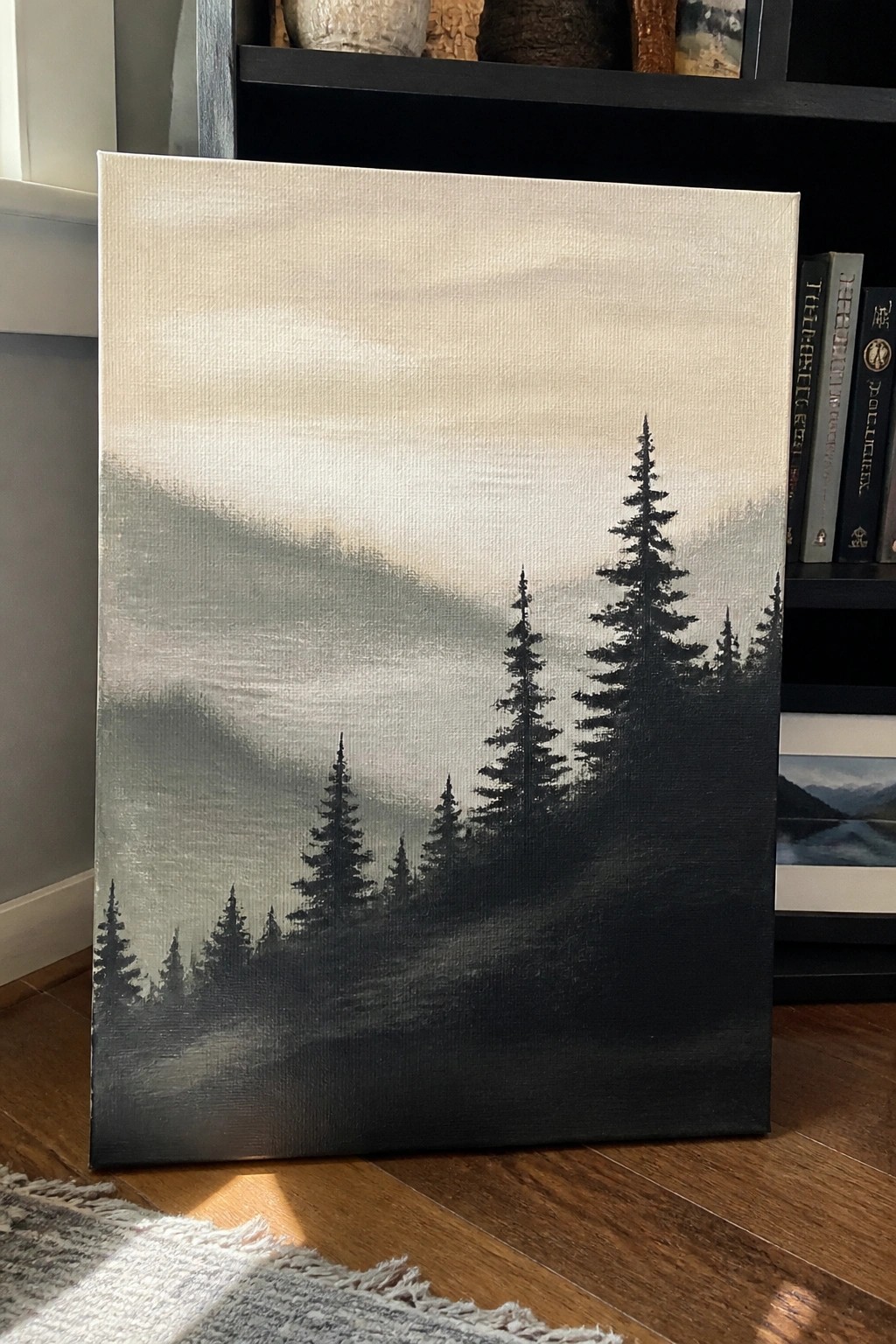Grayscale canvas painting of misty pine trees leaning on bookshelf beside rug