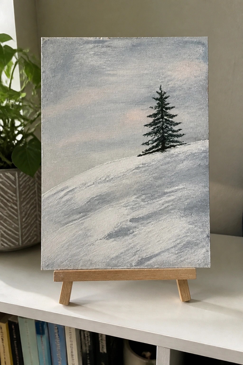 Winter landscape painting of lone pine tree on snowy hill on wooden easel.