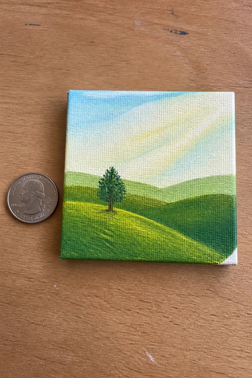 Small canvas landscape of rolling green hills, lone tree, beside quarter coin