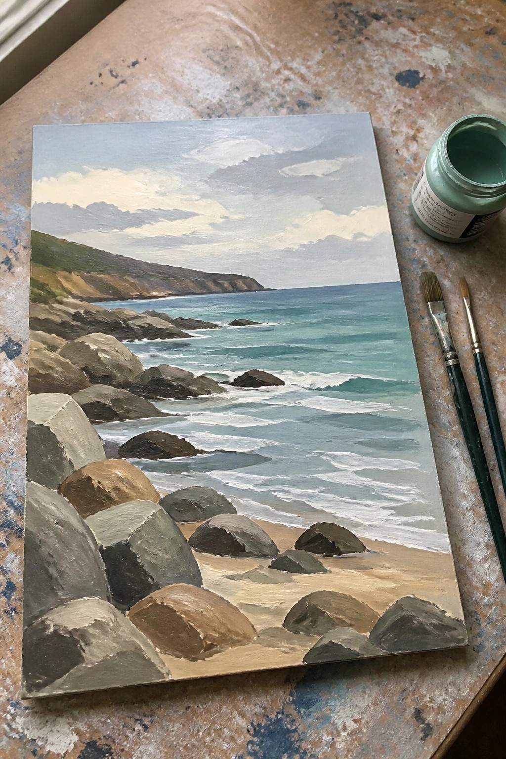 Seascape painting of rocky shoreline with ocean waves beside paint jar and brushes