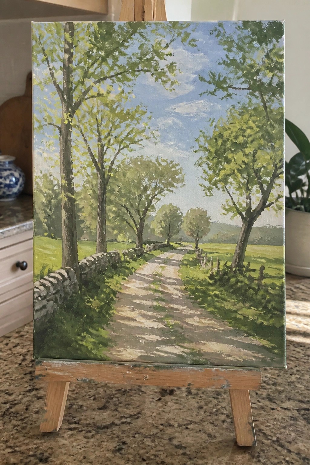 Wooden easel displaying landscape painting of sunny tree-lined dirt path with stone wall