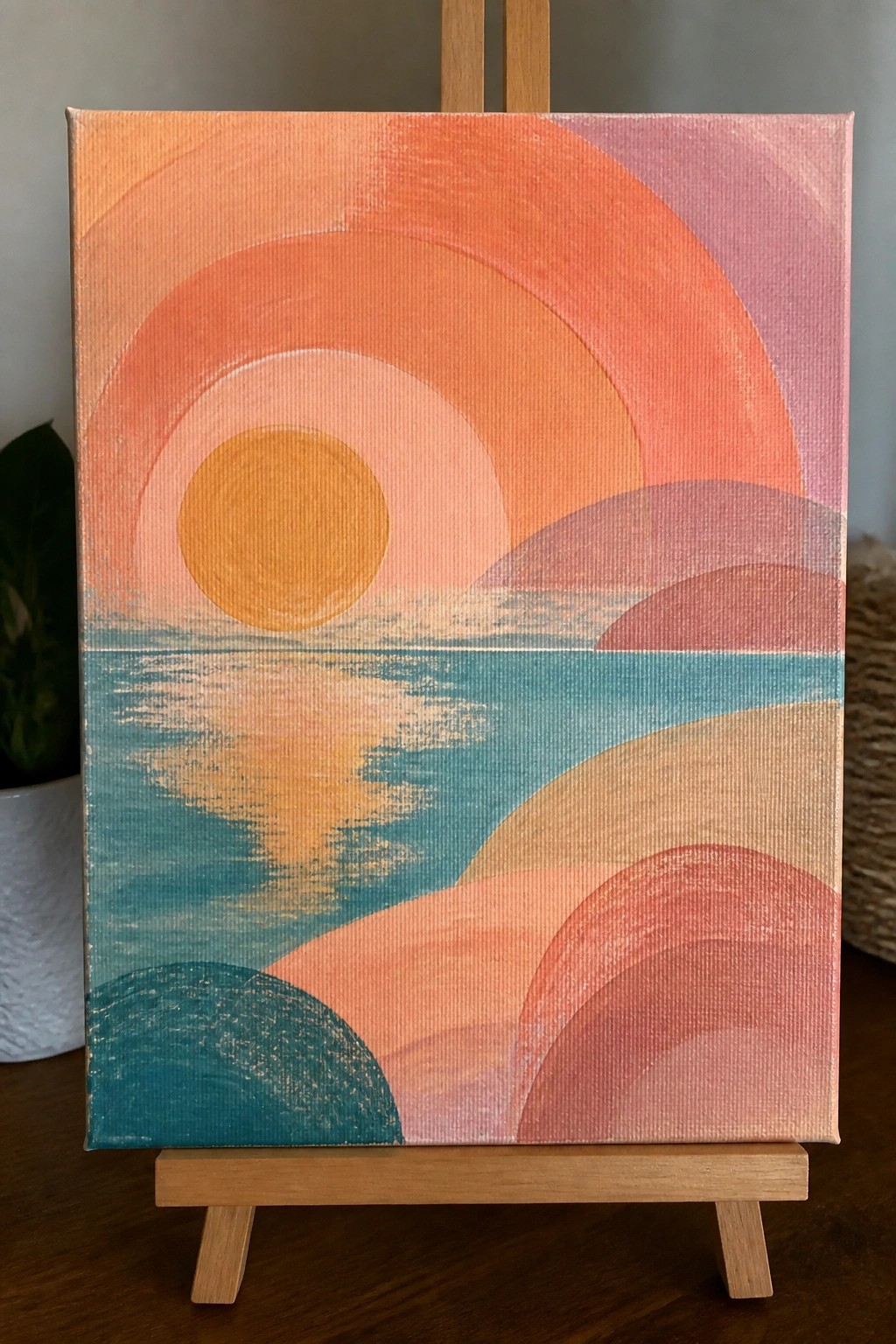 Abstract canvas painting of orange sun over teal water on wooden easel