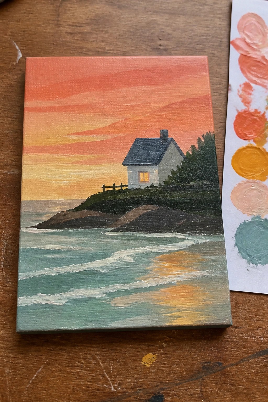 Mini canvas painting of white house by ocean at orange sunset with paint palette nearby