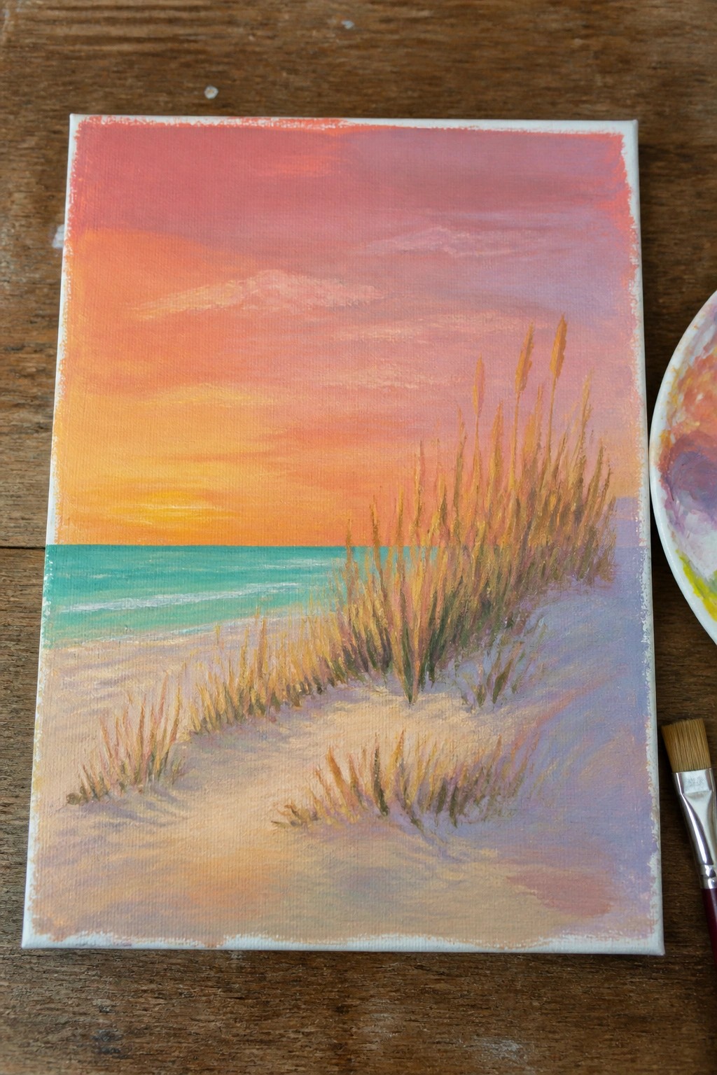 Painting of beach sunset with pink-orange sky, turquoise sea, sand dunes and grass beside palette