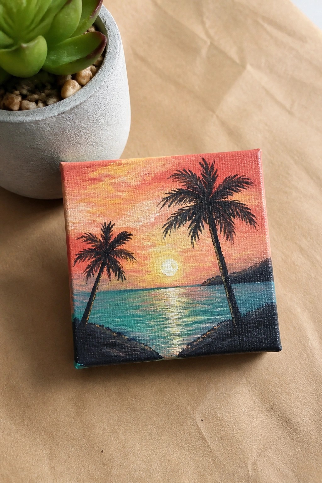Small canvas sunset painting with palm trees beside potted succulent plant