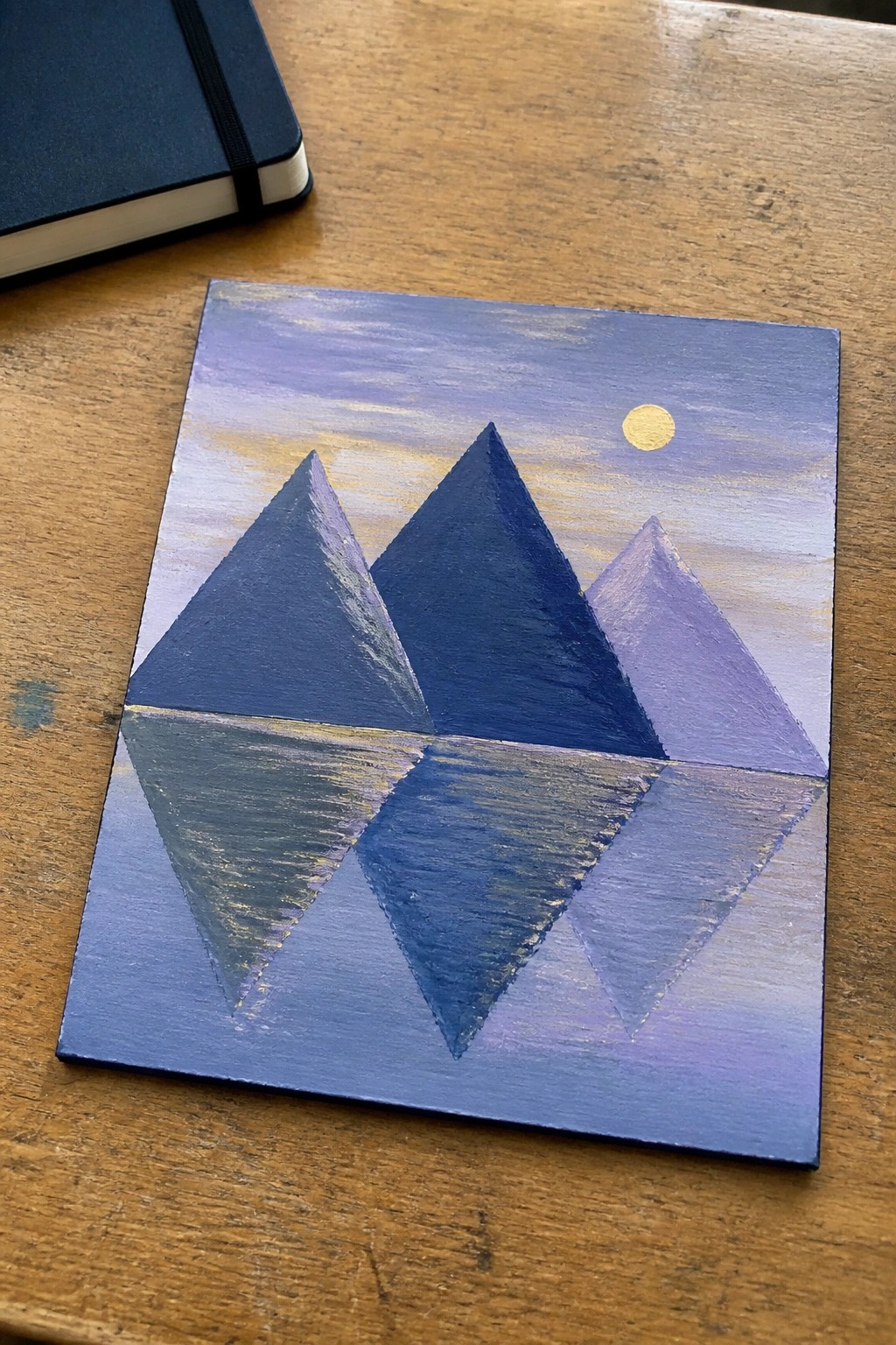 Painting of three blue pyramids reflected below yellow moon on wooden table with notebook