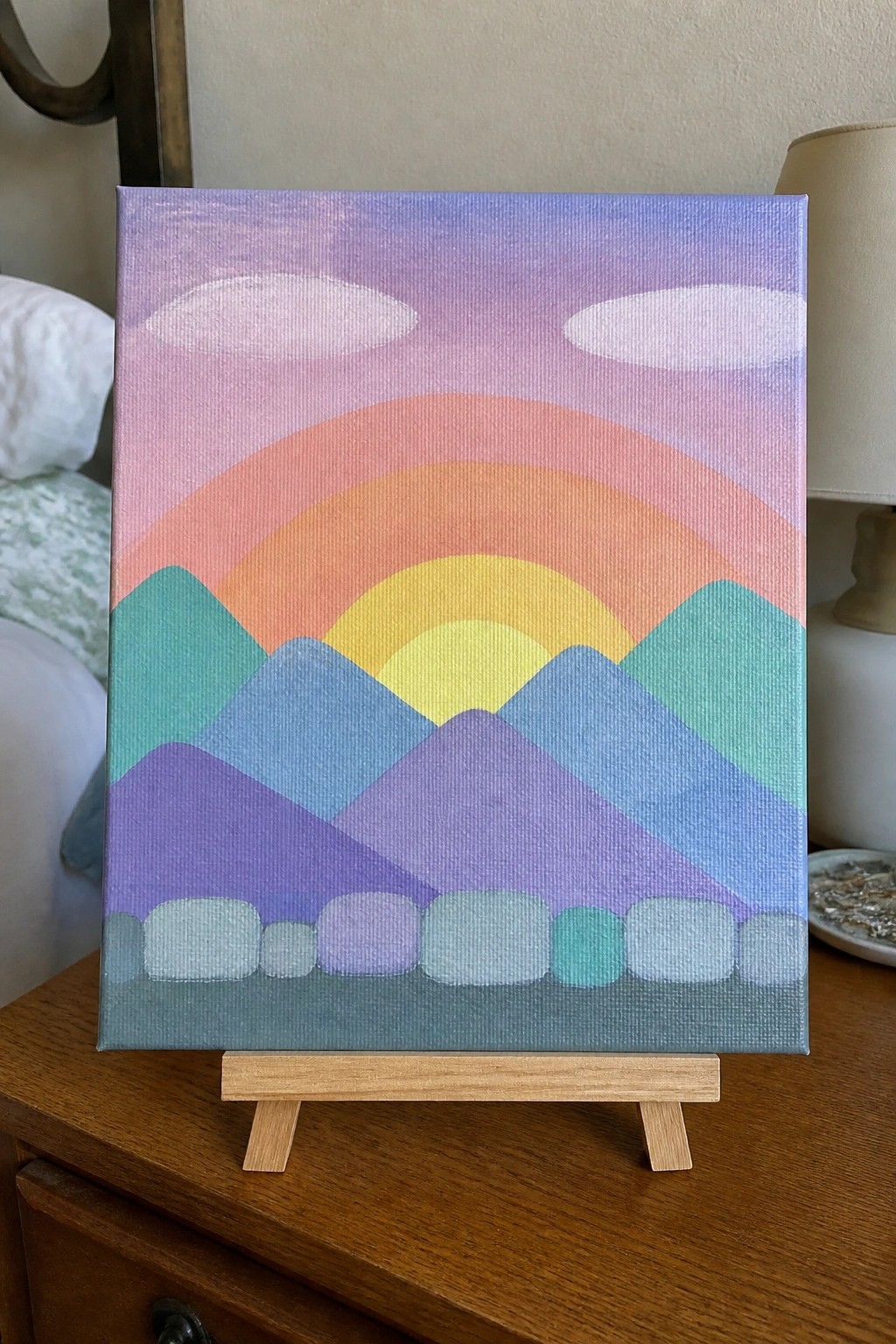 Pastel rainbow arcs over layered purple mountains on canvas atop wooden easel