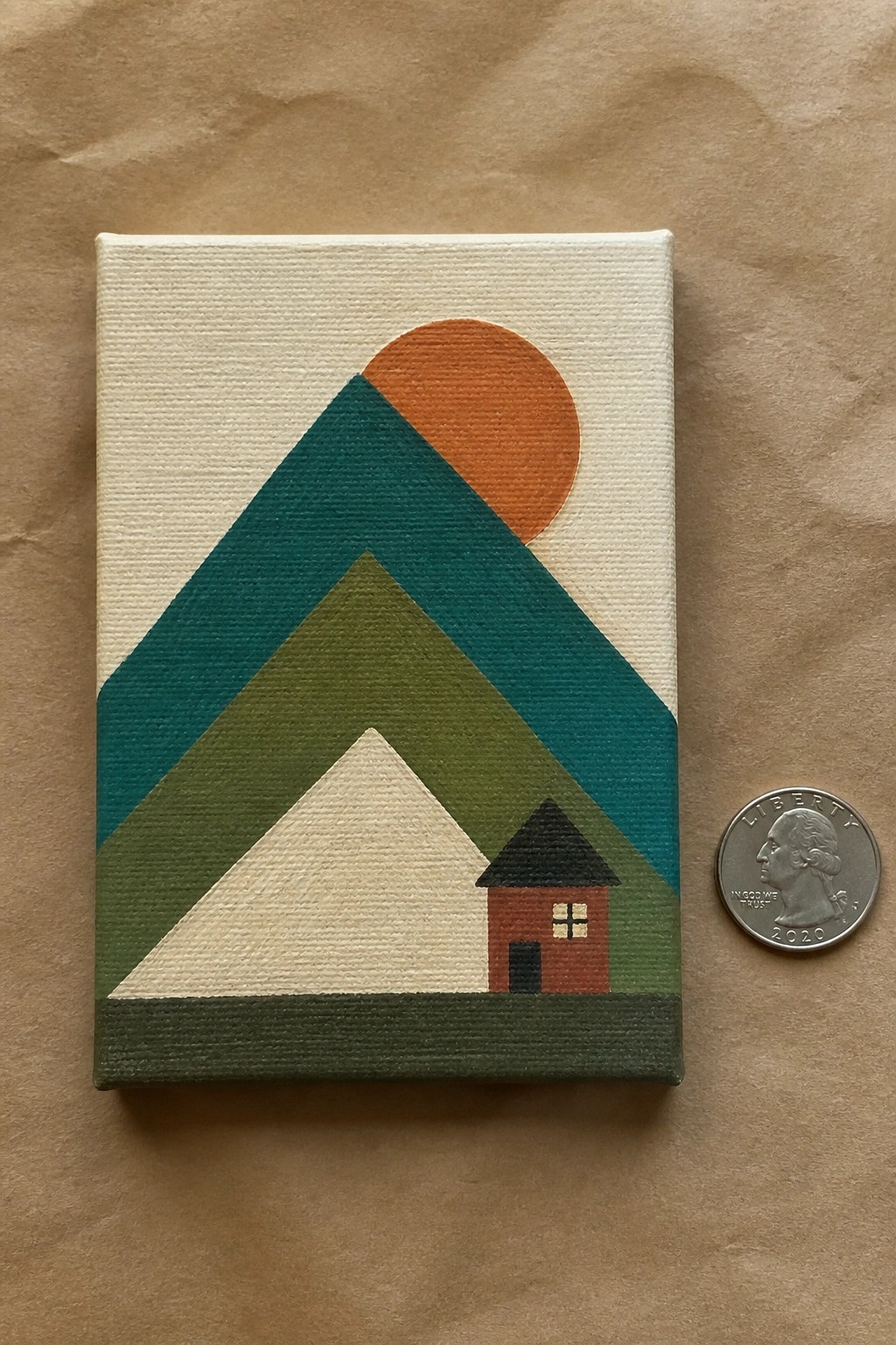 Small acrylic canvas painting of layered teal green and beige triangles forming mountains with an orange sun and red house beside a 2020 quarter on brown paper.