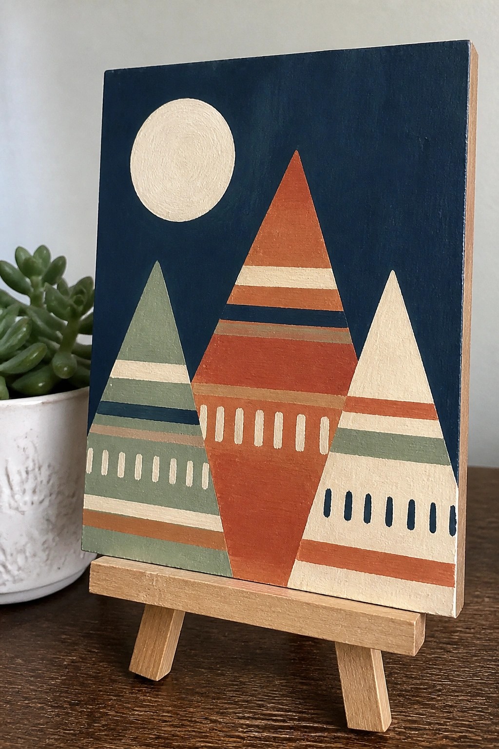 Abstract painting of three striped mountains under moon on wooden easel beside plant