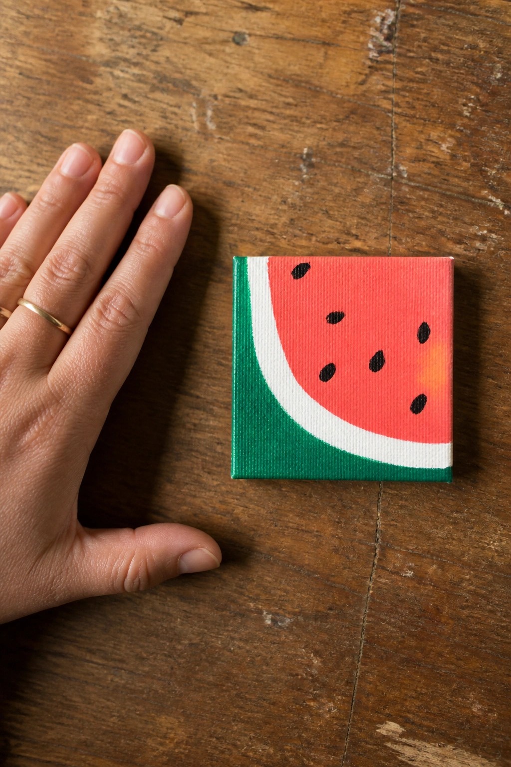 Hand with gold ring beside small square canvas of watermelon slice on wood.