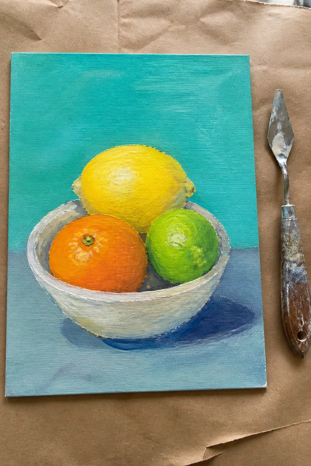Still life painting of lemon, orange, and lime in white bowl beside palette knife