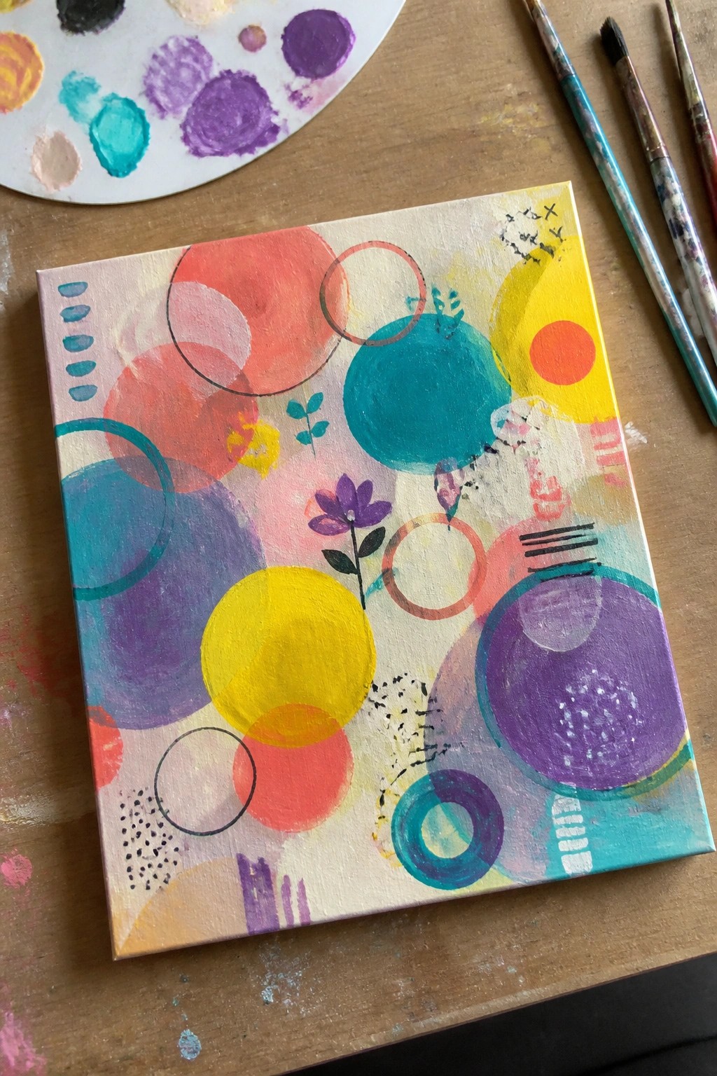 Abstract painting with colorful circles, shapes beside palette and brushes on wood table