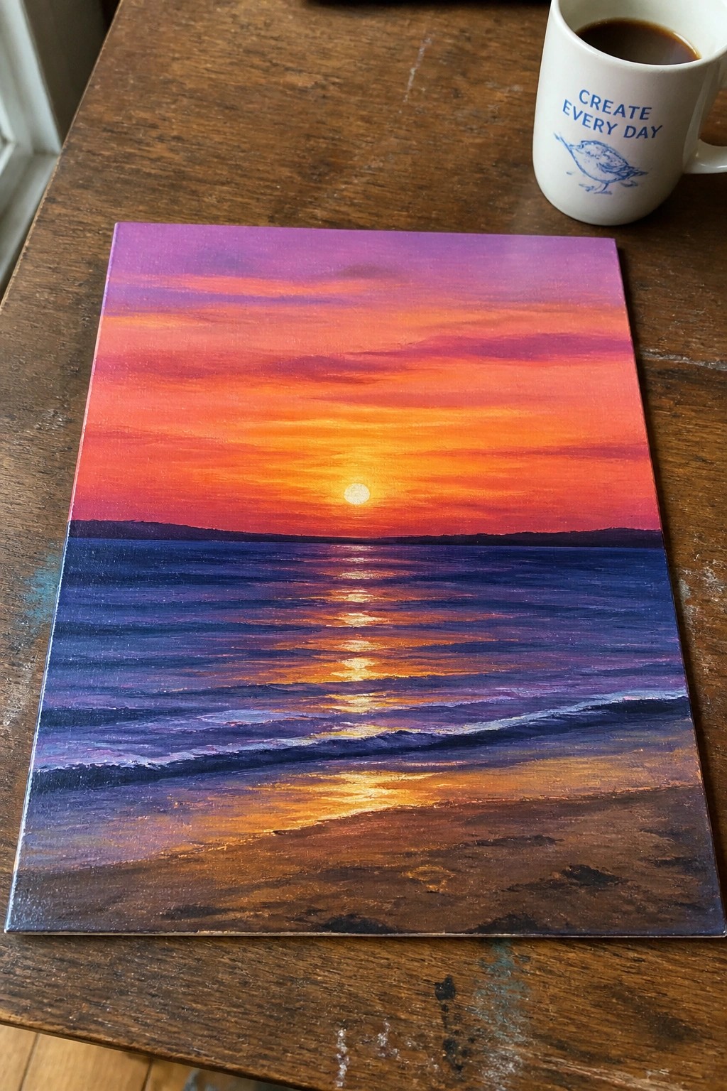Sunset seascape painting with vibrant orange sky beside white coffee mug on wood table