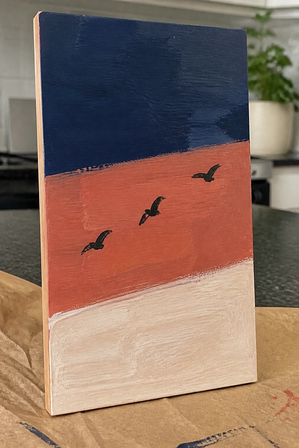 Small wooden painting with blue sky, red band with three black birds, beige base.
