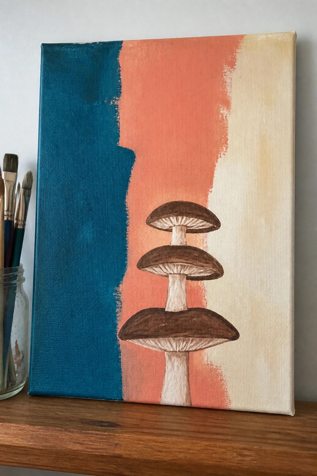 Stacked brown mushrooms on canvas with teal, coral, and beige background beside paintbrushes