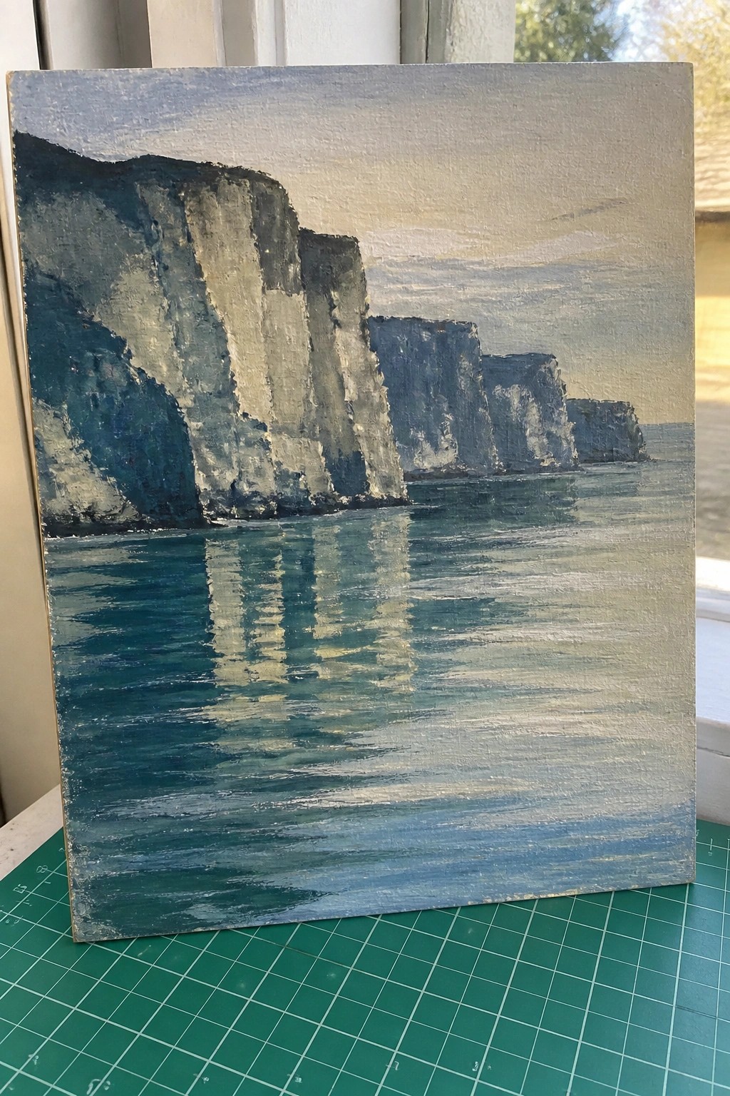 Seascape painting of cliffs reflected in blue water on green cutting mat